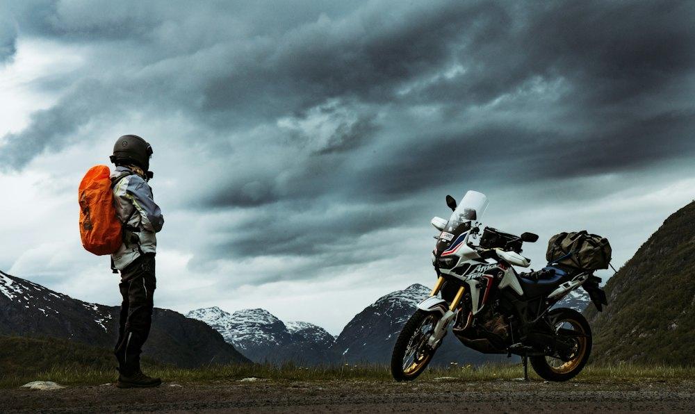 Rider with an orange backpack looking at storm clouds over snowy peaks, her adventure bike parked beside her